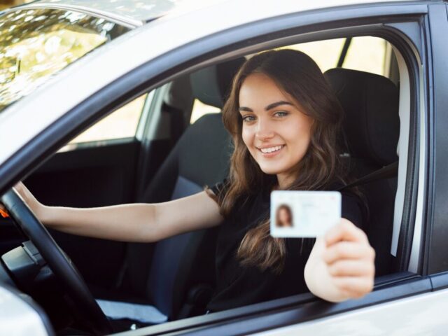 Happy student driver sitting in the modern silver car and showing driving car license to the camera having good mood and happy to pass driving exams